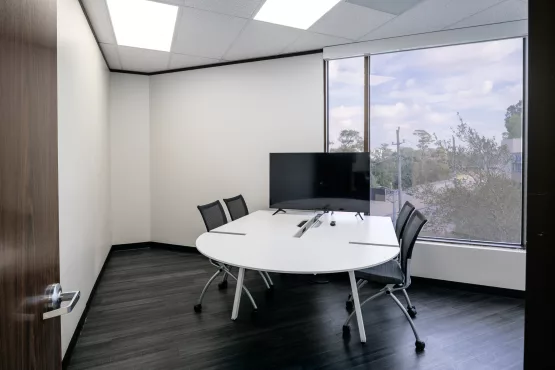 Small meeting room with a round white table, monitor, and large corner windows.
