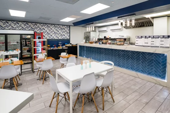 Bright office café with white tables, a service counter, blue tile accents, and a menu board.