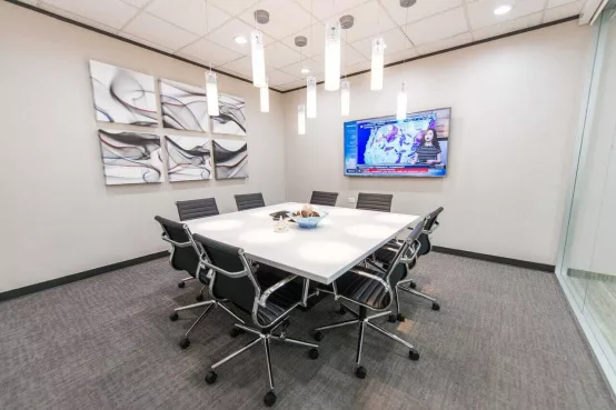 An Avalon Suites conference room with a white square table, modern white chairs, pendant lighting, a wall-mounted whiteboard, a large potted plant, and glass walls opening to the office corridor.