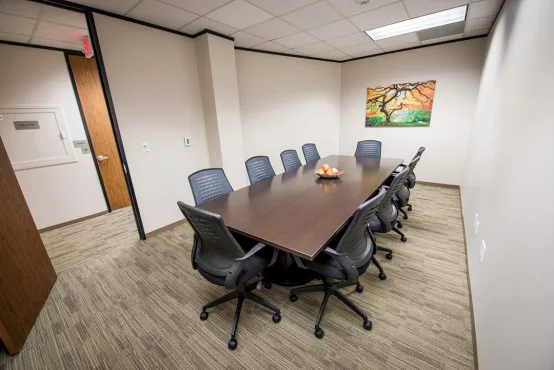 An Avalon Suites conference room with a large dark wood rectangular table, gray mesh chairs, a fruit bowl centerpiece, and a colorful landscape painting on the wall.