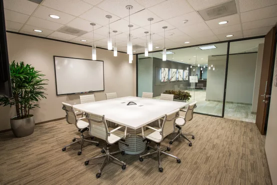 A bright Avalon Suites conference room with a white square table, white chairs, pendant lighting, a whiteboard, and glass walls overlooking the office corridor.