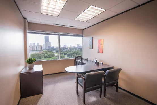 An Avalon Suites private office featuring a dark wood L-shaped desk, two guest chairs, colorful wall art, and panoramic city skyline views through large corner windows.