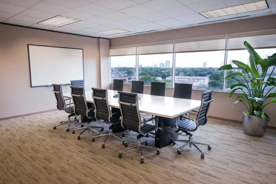 An Avalon Suites conference room with a white rectangular table, black leather chairs, a whiteboard, a large potted plant, and floor-to-ceiling windows offering panoramic city skyline views.
