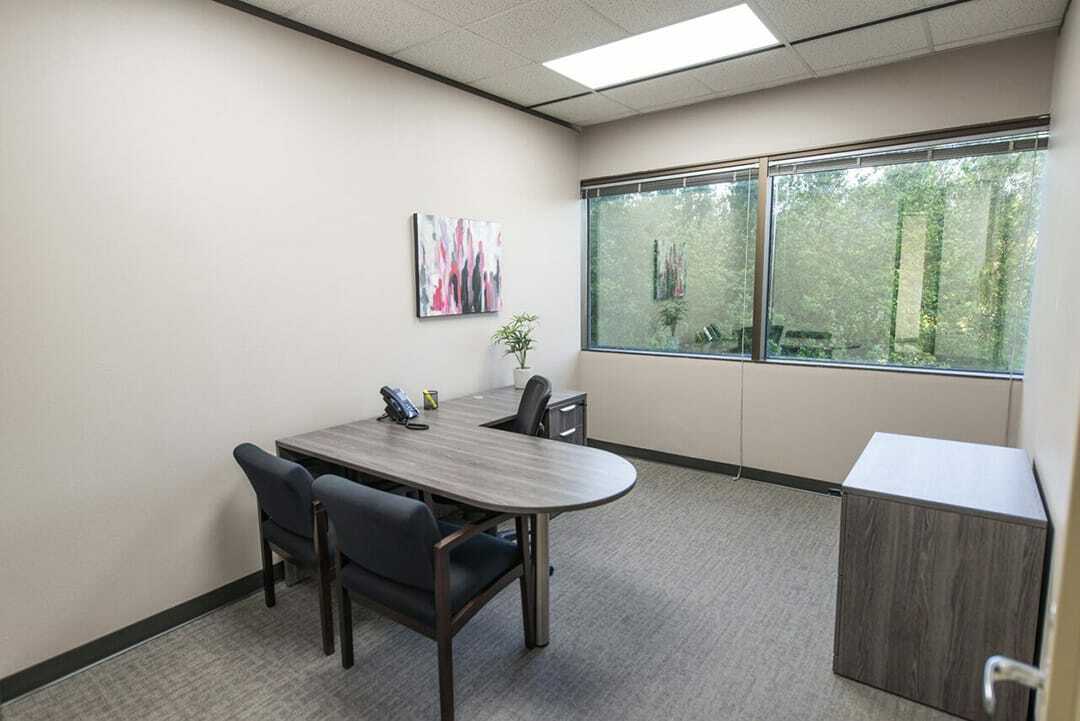 A private office location featuring a rounded wood desk, two guest chairs, an ergonomic office chair, and large windows overlooking lush green trees.